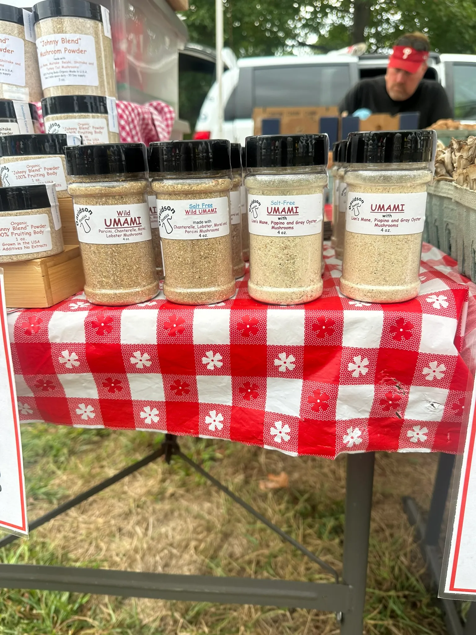 Containers of Johnny Blend powder and UMAMI seasoning shakers lined up on a red gingham cloth at a farmers market table.