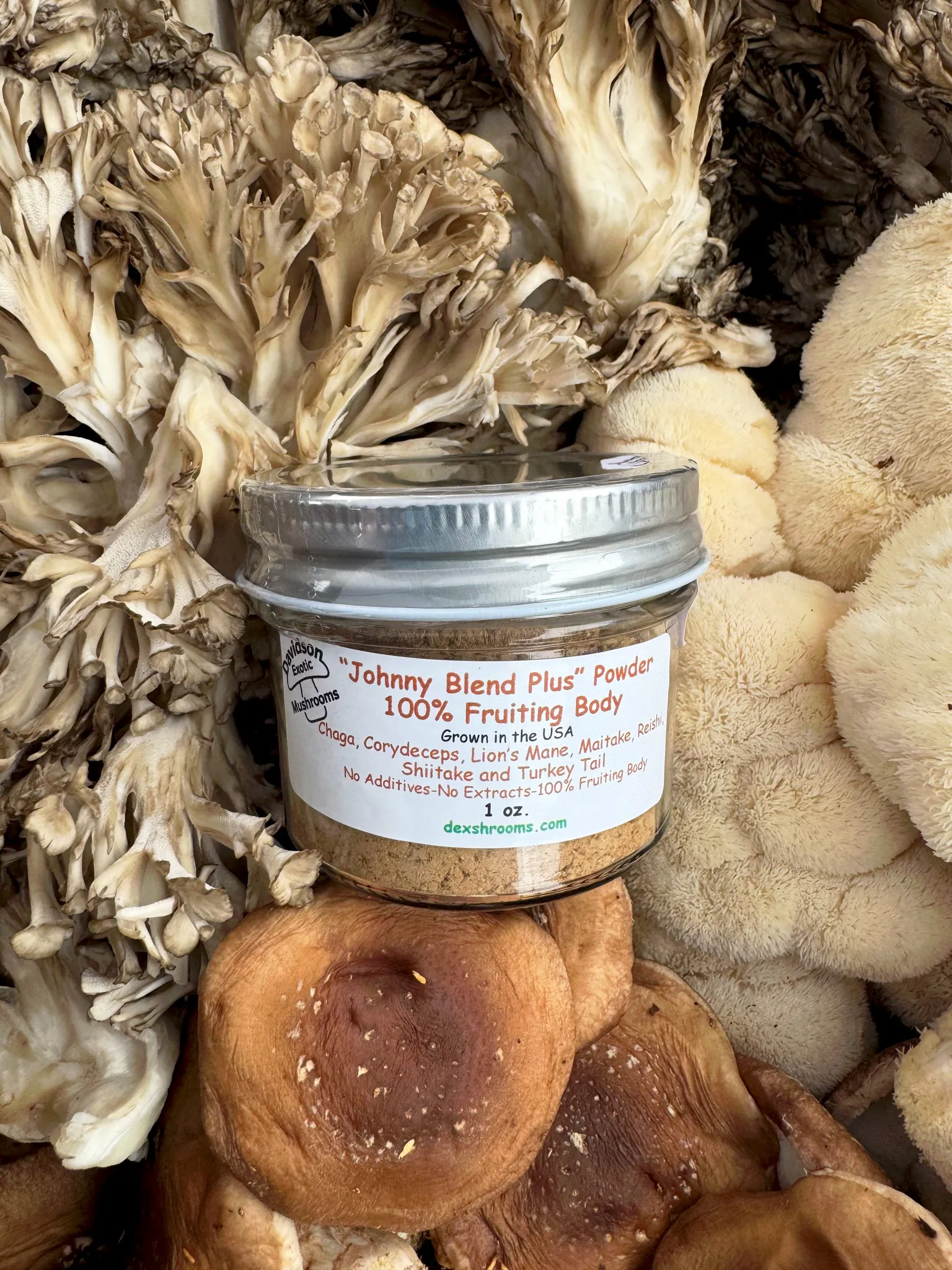 The 1 oz glass container of "Johnny Blend Plus" Mushroom Powder, set on fresh Maitake, Lion's Mane and Shiitake mushrooms.